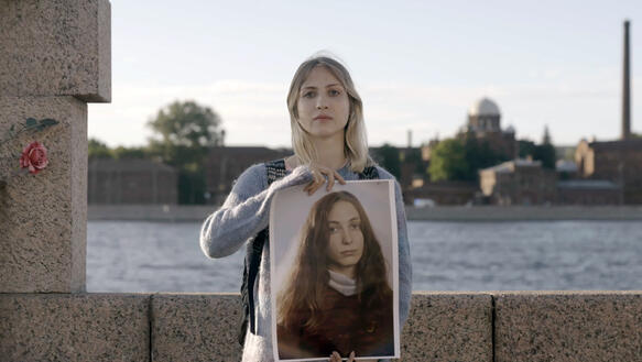Eine Frau mit schulterlangem Jahr steht vor einer Kaimauer, dahinter Wasser, und hält ein Foto-Porträt einer anderen langhaarigen Frau vor ihrem Körper.
