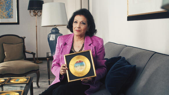 Eine türkische Frau mittleren Alters sitzt in einem Wohnzimmer auf einem Sofa und hält eine gerahmte goldene Schallplatte in den Händen auf ihrem Schoß; sie trägt Halsketten, einen Blazer und ist geschminkt, hinter ihr steht ein Sessel und ein Beistelltisch mit einer Lampe.