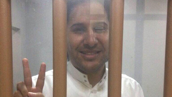 Ein Mann steht hinter Gitterstäben in einer Gefängniszelle, er lächelt und hält die Finger seiner rechten Hand zum Victory-Zeichen gespreizt, er trägt ein Hemd.