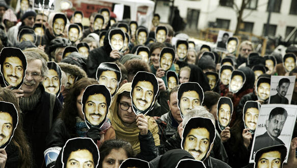 Dutzende Menschen stehen auf einem Platz und halten sich vor ihre Gesicher Masken mit dem Portätfoto des lächelenden Abdulhadi Al-Khawaja.