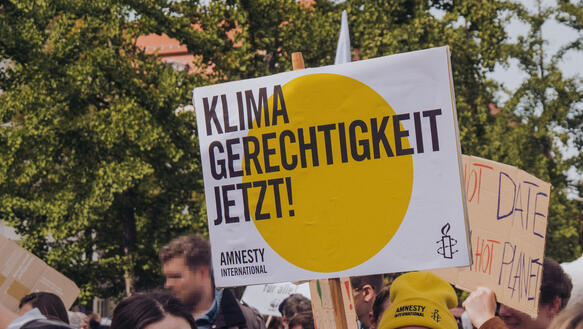 Das Foto zeigt eine dichtgedrängte Menschenmenge auf einer Demonstration. Eine junge Frau mit Amnesty-Mütze hält ein Schild mit Amnesty-Logo hoch. Auf dem Foto steht: "Klimagerechtigkeit jetzt!".