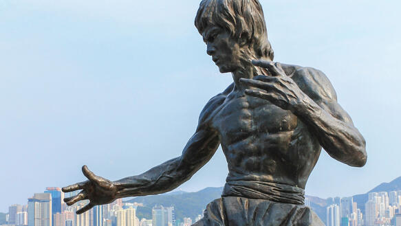 An einem Hafenbecken steht eine metallene Statue des Action-Filmhelden Bruce Lee, im Hintergrund am Ufer die Skyline Hongkongs.