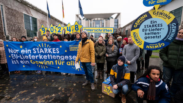 Eine Gruppe von circa 50 Personen steht vor dem Kanzleramt. Einige halten ein großes Banner vor sich. Auf dem Banner steht: "Nur ein starkes EU-Lieferkettengesetz ist ein Gewinn für alle".