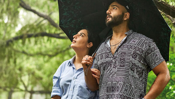 Ein Liebespaar; er hält einen Regenschirm, unter dem beide stehen inmitten eines Parks, sie schauen nach oben in die gleiche Richtung, um sie herum Bäume und über ihnen ein Blätterdach.