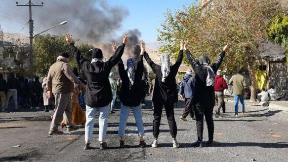 Vier Frauen stehen mit dem Rücken zur Kamera auf einer Straße. Sie heben ihre Hände in die Luft und zeigen mit beiden Händen das Victory-Zeichen. Weitere Menschen demonstrieren auf der Straße. Im Hintergund steigt Rauch auf.