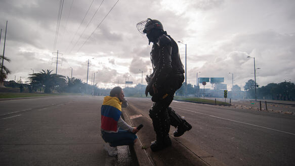 Eine Frau mit kolumbianischer Flagge auf einer Straße in der Hocke, vor ihr ein Polizist.