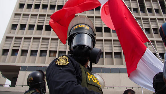 Da Bild zeigt im Vordergrund einen Polizisten mit ABC-Schutzmaske, dahinter die peruanische Flagge