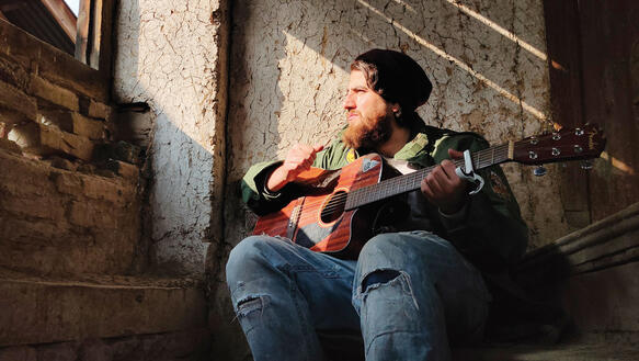 Ein Musiker sitzt mit seiner Gitarre vor einer verwitterten Hauswand auf dem Treppenabsatz zu einem Hauseingang; er trägt zerrissene Jeans, eine Armeejacke und eine Mütze; er hat einen Vollbart und sein langes Haar quillt unter der Mütze hervor.