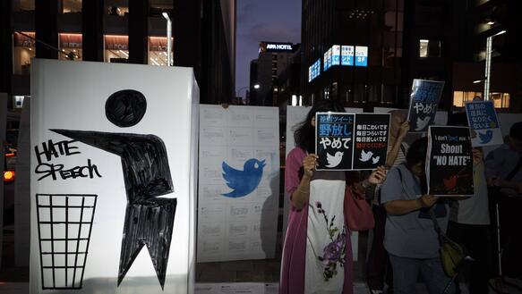 Demonstrant*innen stehen mit Plakaten im Dunkeln auf einer Straße, auf ihren Schildern steht "Shout No Hate" und auf anderen sind japanische Schriftzeichen und das Logo von Twitter abgebildet.