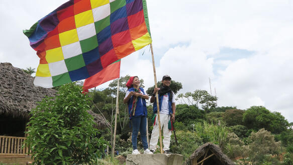 Indigene stehen vor einer Hütte an einem Waldstück und hissen eine Flagge, die im Wind weht; sie lächeln dabei.