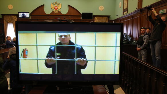 Das Bild zeigt einen TV-Bildschirm, der in einem Gerichtssaal steht. Darauf zu sehen, ein Mann hinter Gittern.