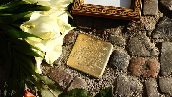 Ein kleiner Gedenkstein aus Messing, in den der Name Paul Otto Hamme eingraviert ist, ist zwischen Kopfsteine in den Boden eingelassen; auf dem Kopfsteinpflaster neben dem Gedenkstein liegen Blumen und ein goldener Bilderrahmen, eingerahmt der Schriftzug "Paul, Hamme".