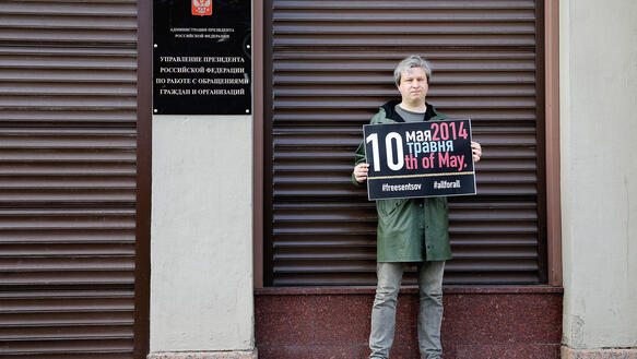 Ein Mann in Jeans, Turnschuhen und Parka steht mit einem Schild (darauf steht "#freesentov, #allforall und verschiedenes in russischer Sprache) vor einem Ladengeschäft, dessen Rolladen heruntergelassen sind.