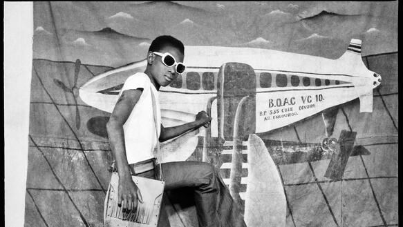 Werbefoto, auf dem ein afrikanisches Model so tut, als steige es in ein Flugzeug, das im Comic-Stil auf eine Leinwand gemalt ist, die an der Wand hängt.