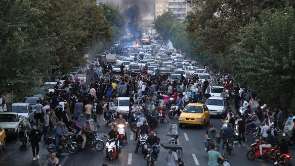 Menschen auf Motorrädern und eine Autoschlange auf einer Straße, im Hintergrund Rauch