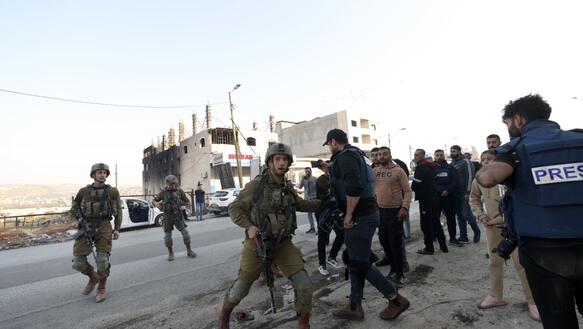 Drei Soldaten mit Helmen und Gewehren stehen auf einer Straße. Einer von ihnen drängt eine Person mit seinem rechten Arm zurück in eine hinter ihm stehende Gruppe.