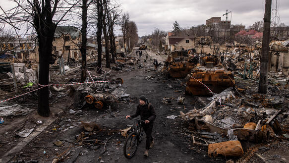 Ein Mann schiebt ein Fahrrad durch einen komplett zerstörten Straßenzug. Fast alle Häuser sind zerstört. Auf der Straße stehen zerstörte und ausgebrannte Panzer.
