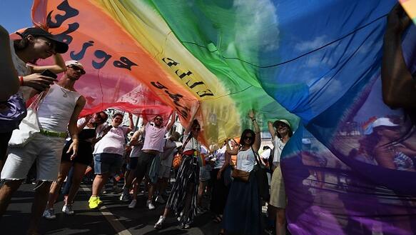 Das Foto zeigt eine Menschenmenge, die gemeinsam eine große und leicht transparente Regenbogenflagge noch oben über ihren Köpfen halten.
