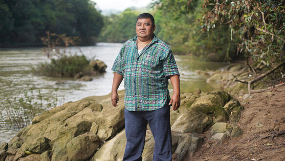 Ein indigener Mann in einem weiten kurzärmeligen Hemd trägt eine lange Hose, die über seine Turnschuhe fällt, seine Arme hängen herunter, während er neben einem Fluss am felsigen Ufer steht, daneben erstreckt sich Wald.
