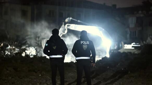 Das Foto zeigt zwei Polizisten von hinten bei Nacht. Sie beobachten einen Bagger, der sich im Scheinwerferlicht durch Trümmer gräbt.