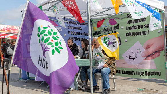 Drei Personen sitzen an einem sonnigen Tag an einem Tisch in einem kleinen Zelt, das mit Fahnen, Schildern und Bannern beschmückt ist. Vor dem Zelt weht eine Fahne im Wind. Auf ihr steht: HDP.