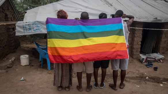 Das Bild zeigt mehrere Personen hinter eine Regenbogenfahne