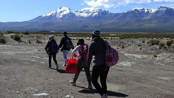 Das Bild zeigt mehrere Personen, die mit Taschen durch eine karge Landschaft gehen, im Hintergrund sind hohe Berge zu sehen.