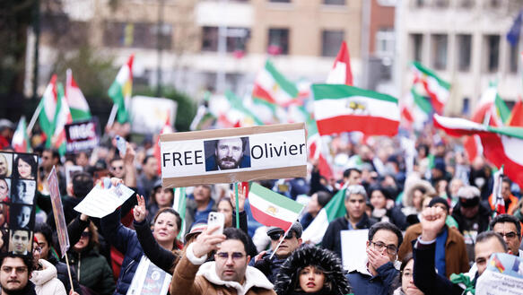 Menschen auf einer Straße protestieren, iranische Flaggen wehen, jemand hält das Schild "Free Olivier" hoch."