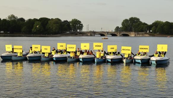 Mehrere kleine Boote treiben nebeneinander auf dem Wasser. Die Personen auf den Booten halten Schilder hoch, die zusammengefügt den Satz ergeben: "Alle Menschen sind frei und gleich an Würde und Rechten geboren".