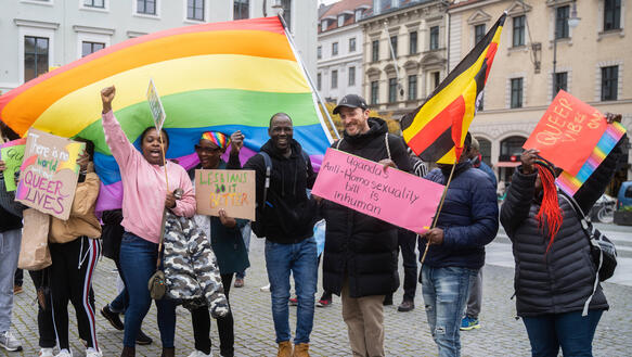 Mehrere Personen stehen auf einem Platz und halten mit Solgans beschriftete Papptafeln hoch. Eine Person schwenkt eine große Regenbogenfahne. Eine andere die Flagge Ugandas.