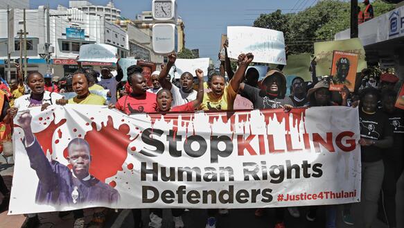 Menschen tragen ein Banner, auf dem steht: „Stop killing human right defenders“.