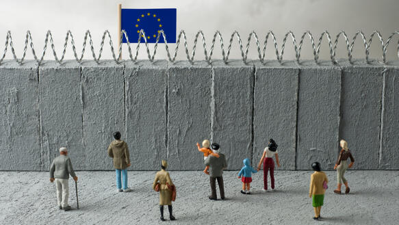 Das Foto zeigt eine Installation mit Spielzeugfiguren. Sie stehen vor einer hohen Mauer, die mit Stacheldraht versehen ist. Dahinter weht die Flagge der Europäischen Union.