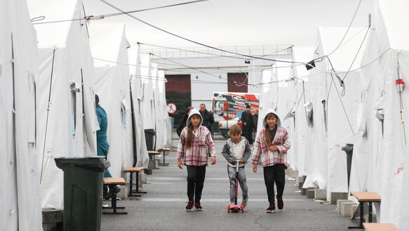Drei Kinder laufen durch ein Flüchtlings-Zeltlager, über ihnen Schnüre, an denen die Zelte aufgespannt sind, hinter ihnen Erwachsene, eins der Kinder fährt auf einem kleinen Roller.