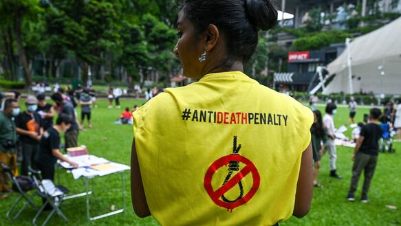 Das Foto zeigt eine von hinten fotografierte junge Frau in einem Park. Auf ihrem T-Shirt steht hinten: Anti deahth penalty. Auf dem T-Shirt ist ein durchgestrichener Galgen abgedruckt.