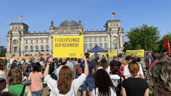 Menschenmenge vor dem Reichstagsgebäude. Eine Person mit langem Haar hält ein gelbes Schild hoch, auf dem "Flüchtlinge aufnehmen!" steht.