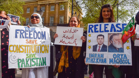 Mehrere Personen stehen vor einem Gebäude und halten Plakate hoch. Auf einem steht: "Release Imran Riaz Khan".