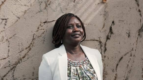Eine mittealte sudanesische Frau in Bluse und Blazer darüber, vor einer Wand.