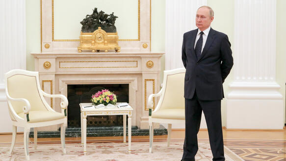 Der russische Staatspräsident Wladimir Putin trägt einen Anzug, hält die Arme hinter seinem Rücken verschränkt und steht in einem barocken Kaminzimmer.