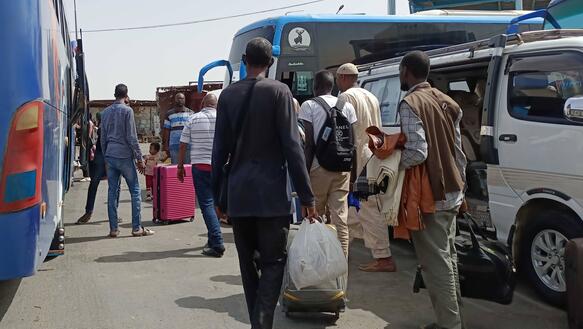 Das Bild zeigt mehrere Personen mit Koffern und Taschen, sie gehen zwischen Bussen und Autos