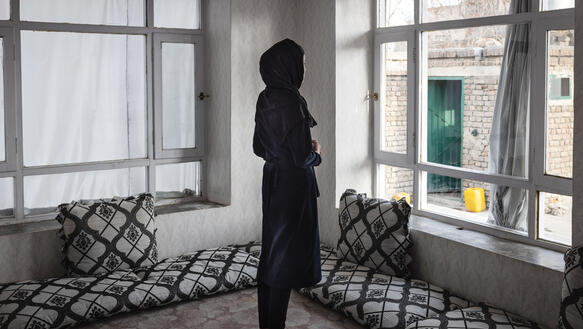 Ein afghanisches Mädchen steht barfuß auf dem Teppichboden in einem Zimmer, hat ein Kopftuch halb über den Kopf gezogen und blickt aus dem Fenster. 
