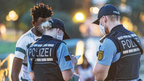 Zwei Polizist*innen, beide tragen Uniform und Mundnasenschutz, kontrollieren einen schwarzen Mann.