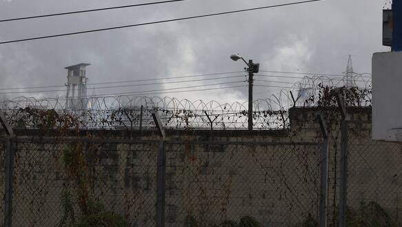 Das Foto zeigt einen mit Stacheldraht versehenen Zaun vor einer Mauer, dahinter stehen Wachtürme.