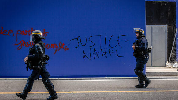 Das Bild zeigt zwei Polizeibeamte vor einer blauen Wand, dort steht geschrieben "Gerechtigkeit für Nahel"
