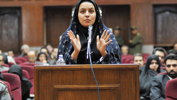 Eine junge Frau mit Kopftuch steht in einem Gerichtssaal vor einem Redepult und gestikuliert mit ihren beiden Händen.