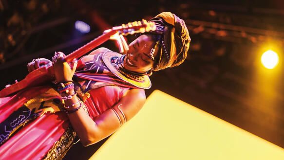 Eine Frau aus Mali musiziert auf einer Bühne, sie trägt traditionelle Kleidung und Schmuck und spielt E-Gitarre.