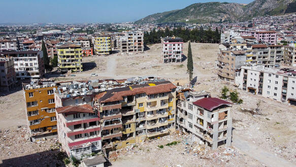 Vogelperspektive auf eine Stadt, deren Häuser von einem Erdbeben schwer zerstört wurden. Dächer sind eingestürzt, Trümmer liegen überall. Am Hoirzont erheben sich Hügel.