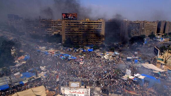 Vogelperspektive auf den Rabaa al-Adawiya-Platz in Kairo: Eine Menschenmenge auf dem Platz während dunkle Rauchschwaden über dem Platz aufsteigen und über die hohen Häuser hinweg in den Himmel steigen. 