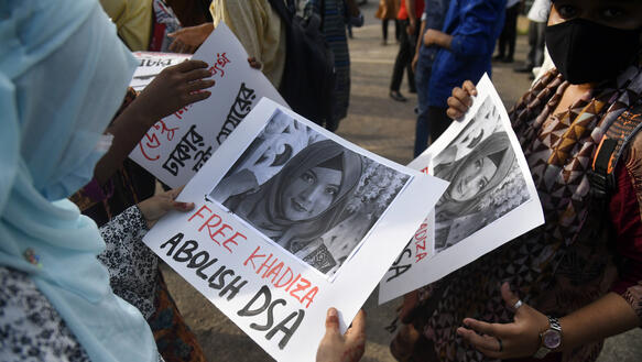 Eine Frau steht in einer Menschenmenge. Sie hält ein Plakat in ihren Händen und betrachtet ist. Auf dem Plakat ist ein Porträtfoto von Khadijatul abgedruckt, auf dem sie ein Kopftuch trägt. Unter dem Foto steht unter anderem: "Free Khadiza".