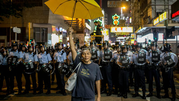 Frau mit gelbem Regenschirm steht in Hongkong vor der Polizei, die die Demonstration auflösen will.