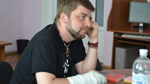 Ein Mann sitzt an einem Tisch und blickt nach rechts aus dem Bild raus. Sein rechter Unterarm und Handgelenk sind bandagiert. Es ist Maksym Butkevych. 